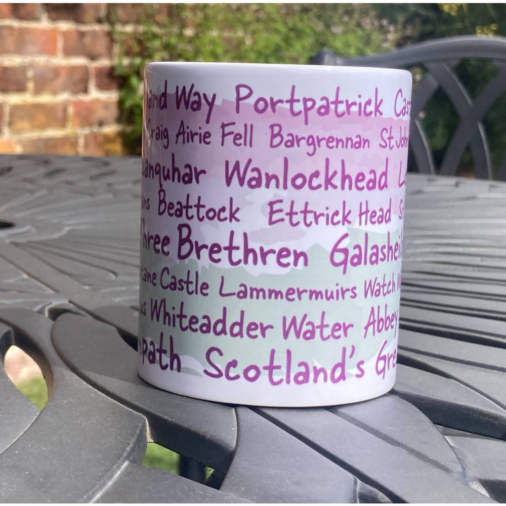 White Southern Upland Way mug with purple text listing locations on the trail a metal table outdoors.