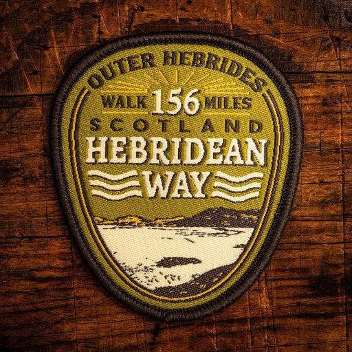 An embroidered shield-shaped patch reading 'Outer Hebrides Walk 156 Miles Scotland Hebridean Way' with a landscape illustration, displayed on a rustic wooden background.