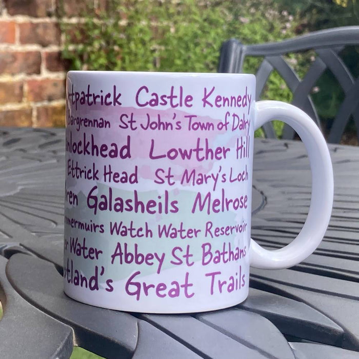 White Southern Upland Way mug with purple text listing locations on the trail a metal table outdoors.