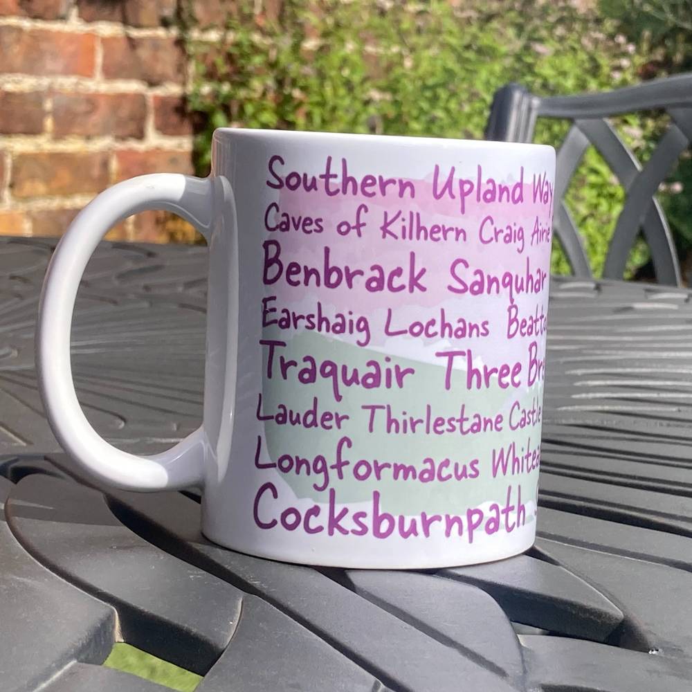 White Southern Upland Way mug with purple text listing locations on the trail a metal table outdoors.
