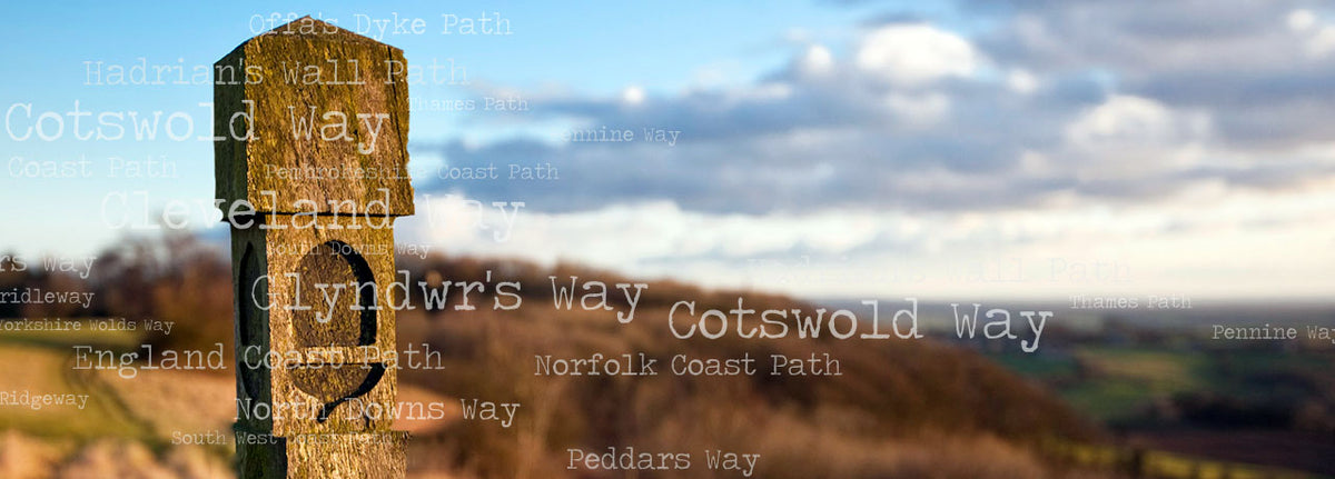 A wooden signpost with the names of National Trails set against a blurred landscape background of rolling hills and a cloudy sky.