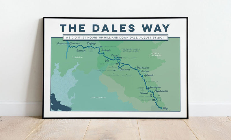 A framed poster print titled 'THE DALES WAY' leaning against a white wall on a light wood floor. The print shows a stylized map in shades of green and blue with route lines and text detailing a 24-hour mountain bike journey.