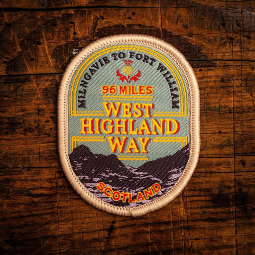 An oval embroidered patch reading 'MILNGAVIE TO FORT WILLIAM 96 MILES WEST HIGHLAND WAY SCOTLAND' with a mountain illustration, placed on a rustic dark wood background.