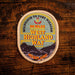 An oval embroidered patch reading 'MILNGAVIE TO FORT WILLIAM 96 MILES WEST HIGHLAND WAY SCOTLAND' with a mountain illustration, placed on a rustic dark wood background.